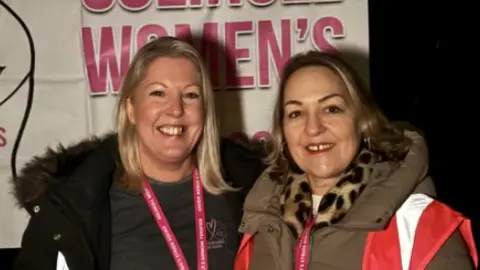 Two women smiling into a camera. They have both got pink high-vis jackets on and coats. They have got pink lanyards on and are smiling. One has blonde hair and one has brown hair. 