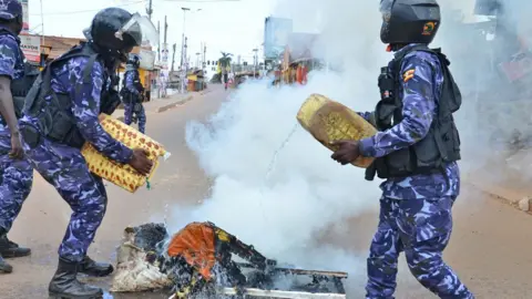 Anadolu/Getty Images Police in blue camouflage and helmets on a Kampala street douse items with water from jerry cans - smoke rises from the pile.