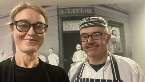 Alexander Taylor A woman wearing glasses, a black cap, long-sleeve t-shirt and apron, and a man with glasses, a white baker's jacket and grey and white striped apron and hat smile towards the camera.