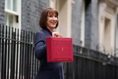 Reuters Rachel Reeves standing in front of black railings holding her red Budget box. She is smiling and wearing a dark blue suit jacket.