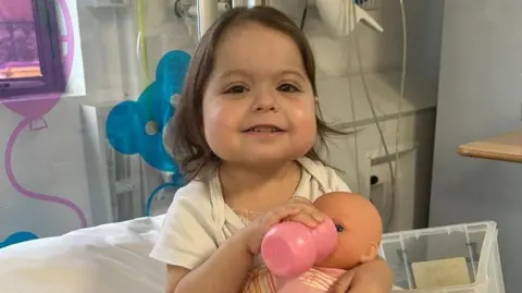 A little girl in a hospital room smiles as she feeds a doll from a bottle. 