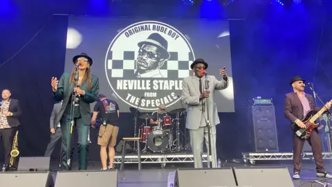 A band perform on a stage. A drummer, guitarist, saxophonist, and two singers can be seen. There is one male and one female singer, both wearing suits and pork pie hats.