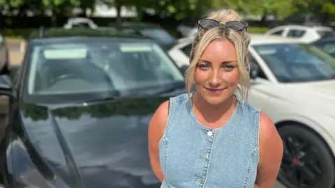 Sophie Rose, who has blonde hair tied up and sunglasses on her head, wearing a denim sleeveless top and standing in front of cars in a car park while looking into camera