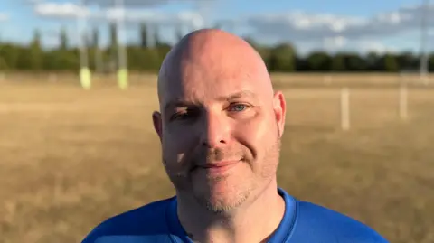Elliot Deady/BBC A man - Matthew Pearson - looking straight at the camera and smiling. He is wearing a blue top and is standing on a rugby pitch.