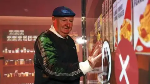 An elderly man who wears a blue cap and green top touches a symbol with a hand sign in a dispclay showing various old cereals. Images of shelves hsowing food jars are in the background.