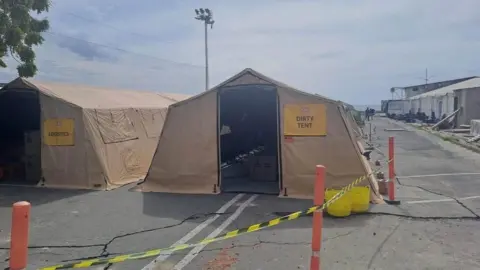 UK International Search and Rescue Two brown tents on grey tarmac. One says "Dirty tent" the other says "logistics". There are orange bollards around them with yellow and black tape connecting them. To the right are more tents. 