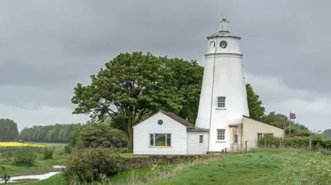 Getty Images A landscape photo of a small white lighthouse with a white shed next to it and a Union flag blowing in the wind behind it. It is surrounded trees and is next to a grass verge and a river.