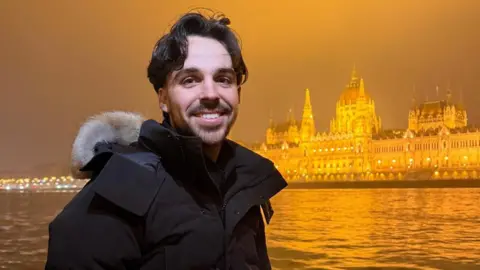 Man with short brown hair and a beard and wearing a black coat is smiling at the camera. Behind him is a river and an ornate building in an orangey/yellow hue.