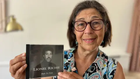 Martin Giles/BBC Angela Young sitting down on a sofa in a living room holding a black and white Lionel Richie CD. She is looking directly at the camera and smiling and is wearing a paisley patterned short sleeve blue blouse.