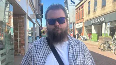 Harvey Bell/BBC A man in a chequered overshirt and white T-shirt standing in a high street. He has reflective sunglasses and a brown haired beard.