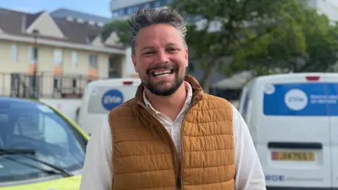 A man stands outdoors in front of parked electric vehicles marked with the EVIe logo, wearing a brown padded vest over a light‑coloured shirt. Residential buildings, trees, and a street are visible in the background.