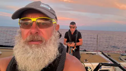 Bob Brown Bob has a big grey beard and is wearing sunglasses on a boat. There is the sea in the background and another man. 