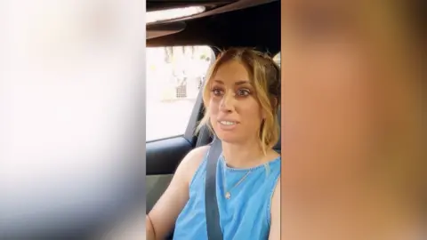A woman with dark blonde hair wearing a blue vest top sits in the front seat of a car.