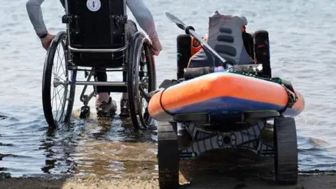 Amy Tallula Will Behenna sat in a wheelchair next to a paddleboard fitted with a special seat and on a trolley rolling down to the water