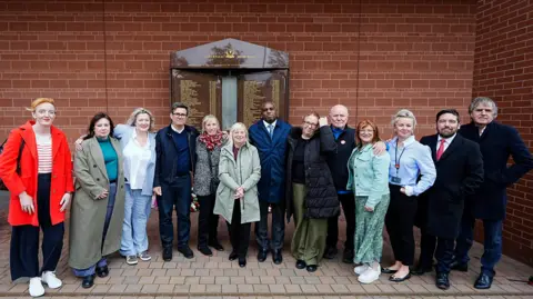 PA Media A group of 13 men and women stand in a line in front of a memorial plaque to the 97 Liverpool fans who were killed at the Hillsborough disaster in 1989. 