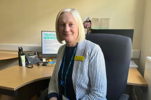 Clare Worden/BBC Carly West-Burnham sits in an office chair in front of her desk. She has a short white blonde bob. She is wearing a white jacket with thin blue stripes. She has a yellow name tag pinned to the right side of her jacket. 
