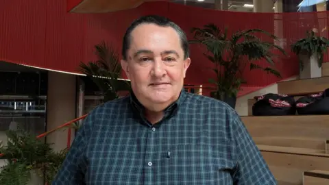 Martin's face is clearly visible and faces forward toward the camera. The hair is short, dark, and neatly combed, with a slightly receding hairline. The face is smooth and clean‑shaven. He is wearing a blue chequered shirt.