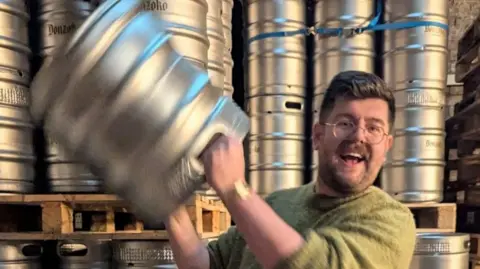 Reece Hugill Reece Hugill smiling at the camera while lifting a metal beer barrel. His arms and the barrel are blurred. He has brown hair which is swept to the side and is wearing a green jumper with the sleeves rolled up showing a gold watch which is also blurred. There are stacks of barrels behind him.