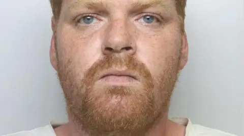 Close up of a mugshot of a ginger man with a beard and blue eyes.