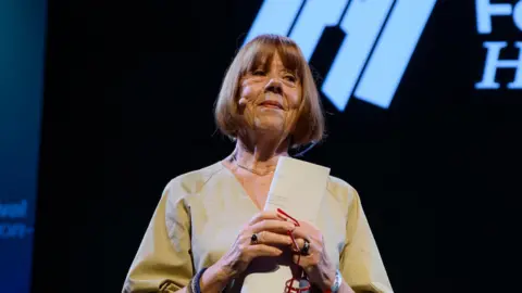 PA Media Gisele on stage at the hay festival. she is wearing a loose fitting, v neck beige top and has red hair in bob style. she is clasping her hands in front of her chest, holding a folded piece or paper and red rimmed glasses. 