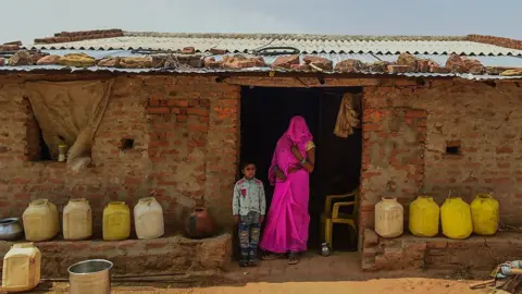 Getty Images A woman in a bright pink saree with her head covered and a child wait for a government water tanker outside their house in Madhya Pradesh, India. Large parts of North India experienced a drought earlier this year. 