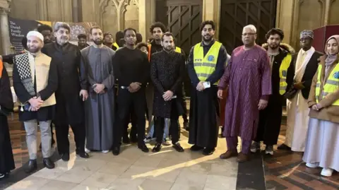 A group of men stand together in a group smiling at the camera. The men are dressed in religious dress for ramadan. The Cathedral walls are seen behind the group as they stand within the entrance. 