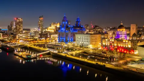 A night city scape of Liverpool lit up in different colours looking out over the docks.