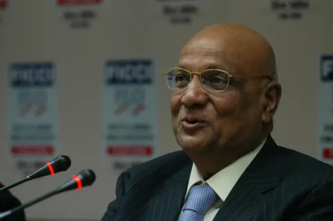 Dijeshwar Singh/ Hindustan Times via Getty Images A close-up of a man wearing glasses, a white shirt,  a blue tie and a pin-striped suit. He sits in front of two small microphones.