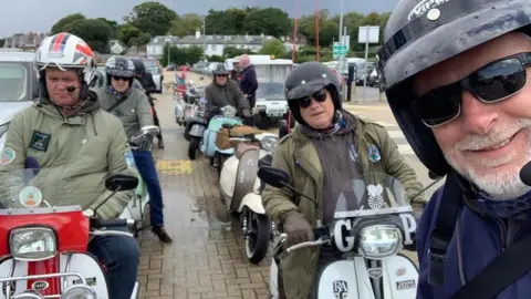 The image is a selfie taken by a man in a helmet, it shows him smiling at the camera and behind him are a group of men on scooters in two lines parked one behind the other on a road. 