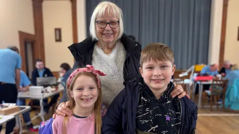 A lady with white hair in a white jumper with two children. The girl is wearing a pink top and pink bow in her hair and the boy is wearing a black and green top and coat. They are all smiling.