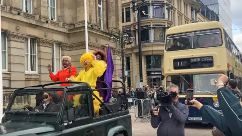 a black land rover carrying three people who are standing up with their arms in the air. One is wearing yellow, with a yellow wig, another is wearing a red outfit, and the other is wearing a purple outfit. There is a double decker bus behind them.