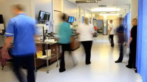 Peter Byrne/PA Wire Busy NHS staff in a brightly lit hospital corridor, with blue curtains to the right and monitors and other equipment to the left.