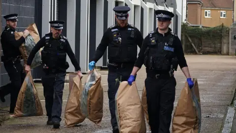 Police leave a unit in Fort Road, Eastbourne with brown bags in each hand. The officers are wearing uniform and two are wearing gloves. The brown bags are marked "evidence" and a transparent section shows plants inside. There is a row of units behind them.