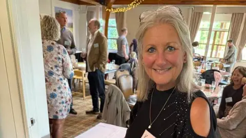Kim Cotton A woman with long grey hair is smiling to the camera. She has reading glasses on her head and is wearing a dark blue dress. She is standing in a hotel conference room with people behind her. 