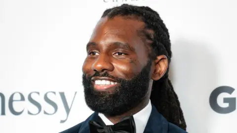 Getty Images Wretch 32 at the GRM Gala. Wretch has long braided hair tied back and a short beard. He smiles, looking to the left of the camera, while wearing a black suit with velvet bow tie