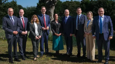 State of Guernsey Six white middle-aged men in dark blue or navy suits and three smartly dressed women standing together in a garden. They are all looking at the camera smiling. There are trees in the background and blue skies. 