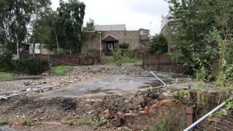 A small rundown brick building is surrounded by waste ground and overgrown bushes.