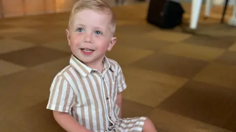 Family Hudson is wearing a brown and white stripy top and shorts and is sitting on the floor smiling at the camera
