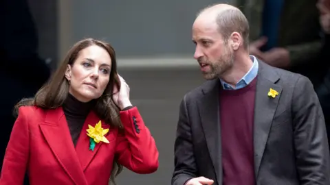 Prince WIlliam and Princess Catherine walking through Wales in the run up to St David's Day hence the daffodil's on their jackets. William is wearing a maroon sweater under a jacket. Catherine is wearing a red jacket.