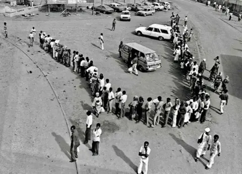 Sunmi Smart-Cole A photo by Sunmi Smart-Cole entitled: "Why... No Buses" - 1984, showing Nigerians waiting in a line for a bus