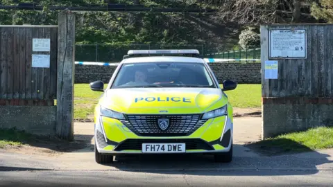 Jenny Kirk/BBC A marked fluorescent yellow and blue police car is parked outside of one of the entrances to Sparrow's Nest Gardens. Behind the car, which is parked, there is a blue and white police tape. 
