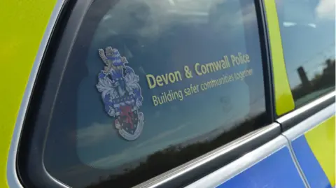 A picture of the Devon and Cornwall police logo on the side window of a police car. The car is bright yellow and blue.