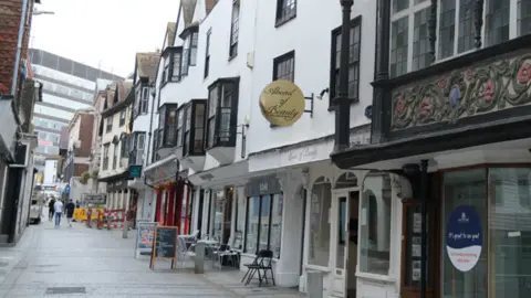 A narrow shopping street with a mix of shopfronts. Image is taken from outside on of the shops looking up the lane