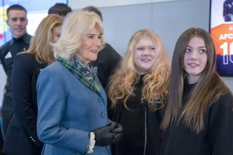 PA Media Queen Camilla, wearing a light blue jacket and a flash of tartan, smiling, with two young women.