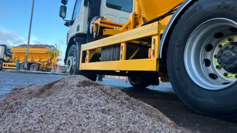 The picture shows a small pile of salt next to a yellow gritter machine. The tyres are large in the frame, while another gritter is in the background. 