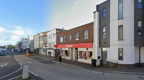 Brewery Lane has a Ladbrookes betting shop and a Coop along it. There are no cars moving cars but two are parked along the pavement. Opposite them is a skip parked next to a bus stop.