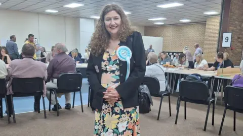 Caroline Gladwin at the count. People are sat at desks behind her. She wears a Reform rosette, and a dark blazer over a flowery dress. She has long curly light brown hair.
