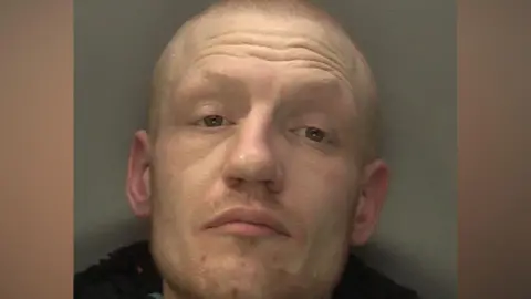 Reece Allen pictured in a mugshot with a neutral expression on his face. He is bald and stood against a grey background