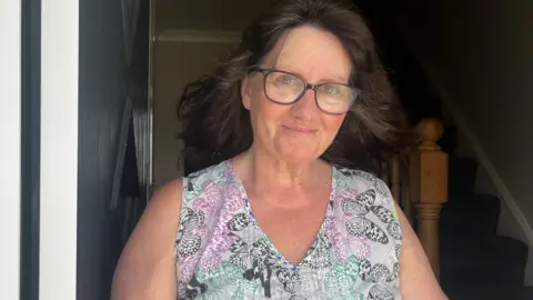 Nicola Haseler/BBC A woman with glasses and wavy brown hair smiles at the camera from her doorway.