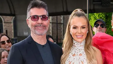 Getty Images A man in dark glasses and a black suit and grey t-shirt smiles next to a woman with long blonde hair in a half-up style, a diamante lacework sleeveless high neck top and diamond earrings. Onlookers stand behind them at a red carpet event.
